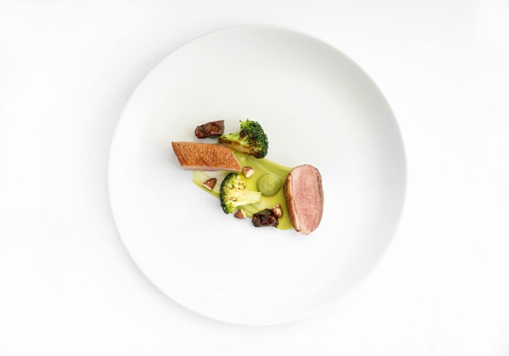 Minimalist gourmet dish with duck, broccoli, and sauce on white plate.