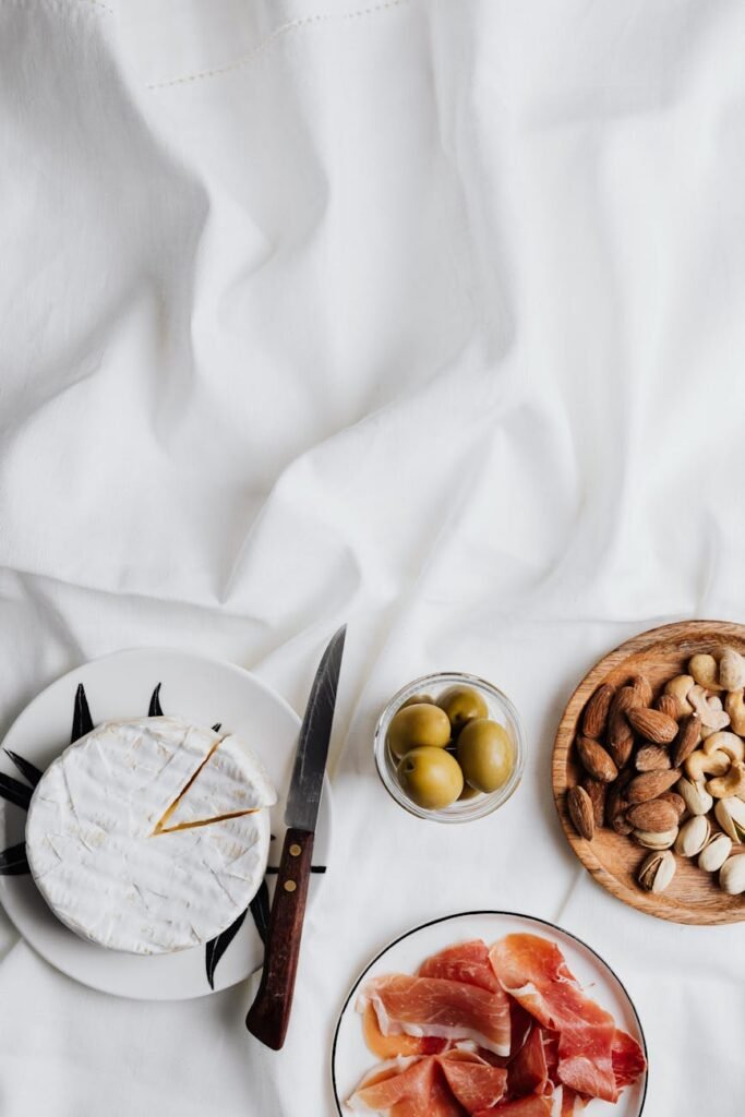 pexels-photo-5202234-5202234 A top view of a charcuterie spread featuring camembert, prosciutto, olives, and nuts on a white background.